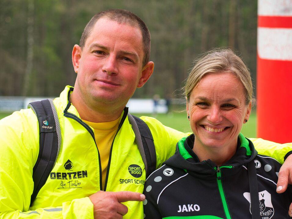 Zwei Personen in Sportkleidung lächeln vor einer roten Fahne, im Hintergrund sind Bäume sichtbar.