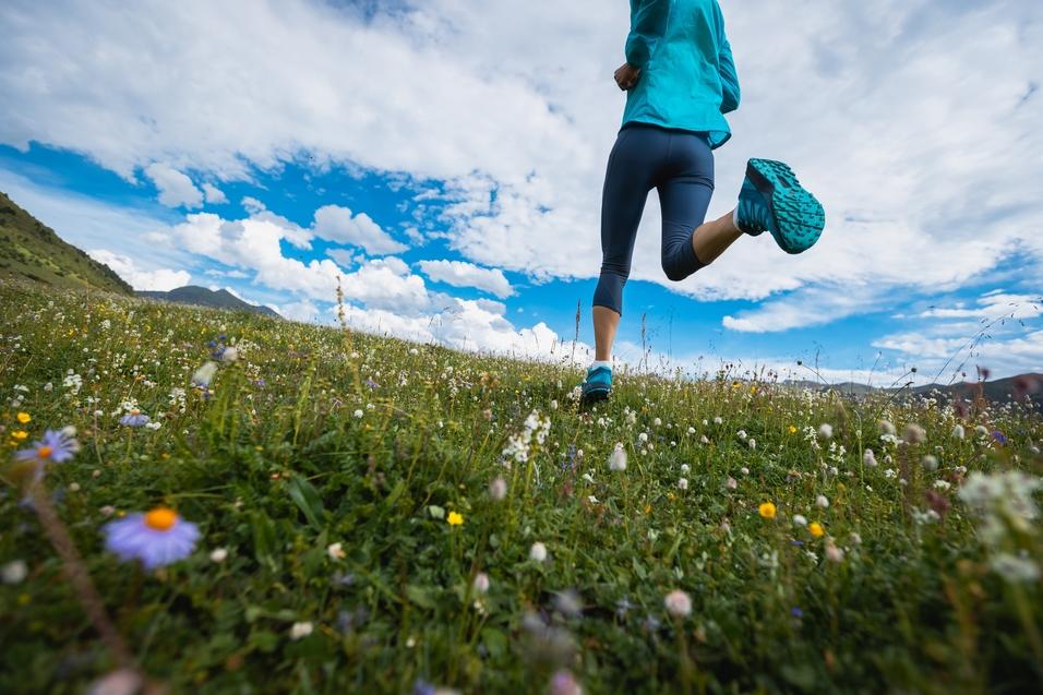 L&auml;uferin in blauer Sportbekleidung joggt &uuml;ber eine bl&uuml;hende Wiese unter einem wolkigen Himmel.