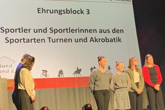 Ehrungsblock mit Sportlern aus Turnen und Akrobatik auf B&uuml;hne mit Pr&auml;sentationswand im Hintergrund.