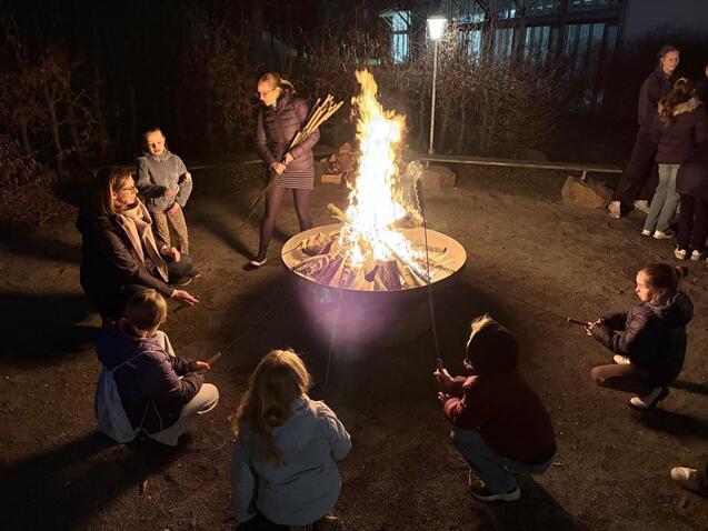 Gruppensitzung um ein Lagerfeuer, Kinder und Erwachsene mit Stockbrot, beleuchtet von Flammen in n&auml;chtlicher Umgebung.
