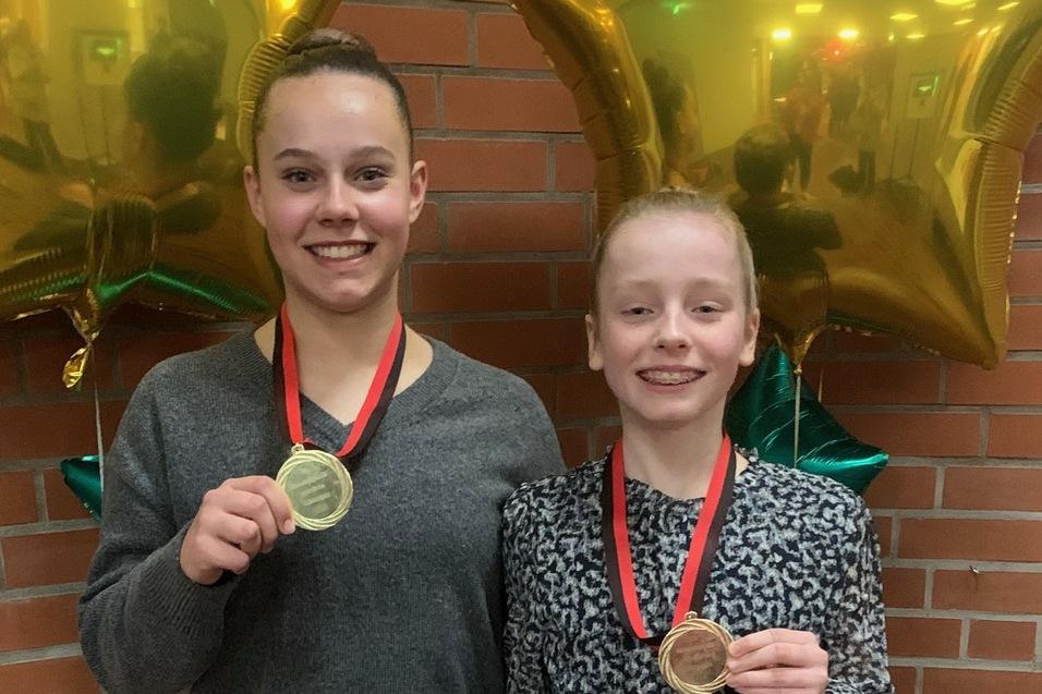 Zwei Mädchen stehen lächelnd mit Medaillen um den Hals vor goldenen Sternballons in einem festlichen Raum.