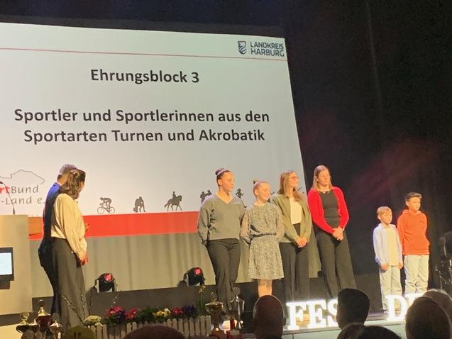 Ehrungsblock mit Sportlern aus Turnen und Akrobatik auf B&uuml;hne mit Pr&auml;sentationswand im Hintergrund.