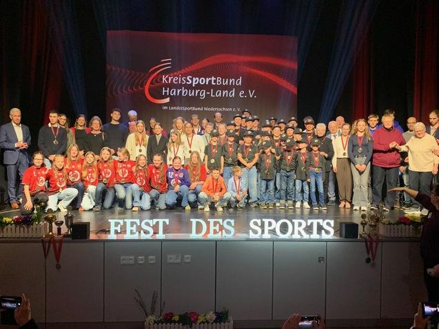 Gruppenzusammenkunft von SportlerInnen und Trainern beim Fest des Sports mit Medaillen und Troph&auml;en im Hintergrund.