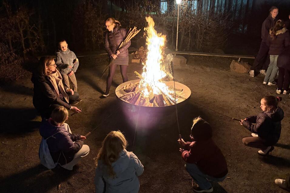 Gruppensitzung um ein Lagerfeuer, Kinder und Erwachsene mit Stockbrot, beleuchtet von Flammen in n&auml;chtlicher Umgebung.