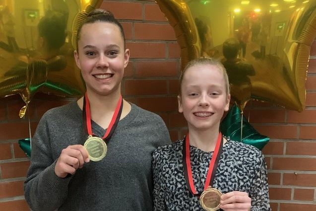 Zwei M&auml;dchen stehen l&auml;chelnd mit Medaillen um den Hals vor goldenen Sternballons in einem festlichen Raum.