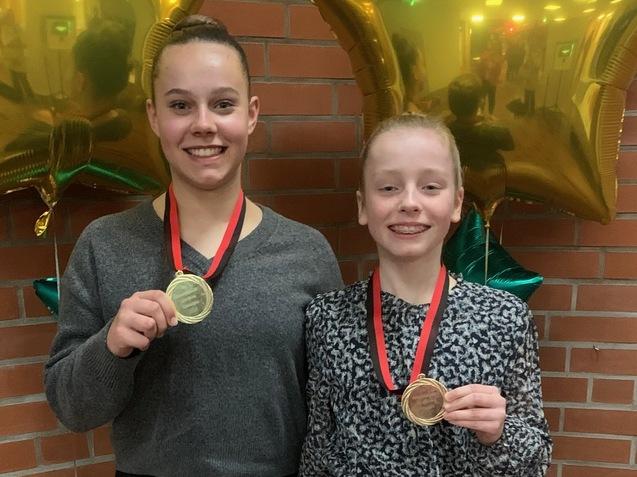 Zwei M&auml;dchen stehen l&auml;chelnd mit Medaillen um den Hals vor goldenen Sternballons in einem festlichen Raum.