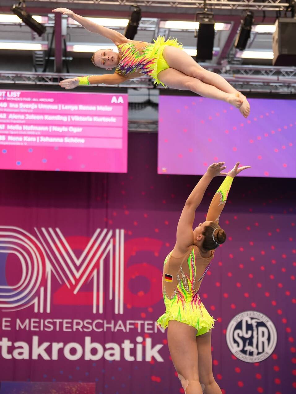 Zwei Turnerinnen f&uuml;hren einen Sprung im Sportakrobatik-Wettbewerb vor einer bunten Kulisse aus.