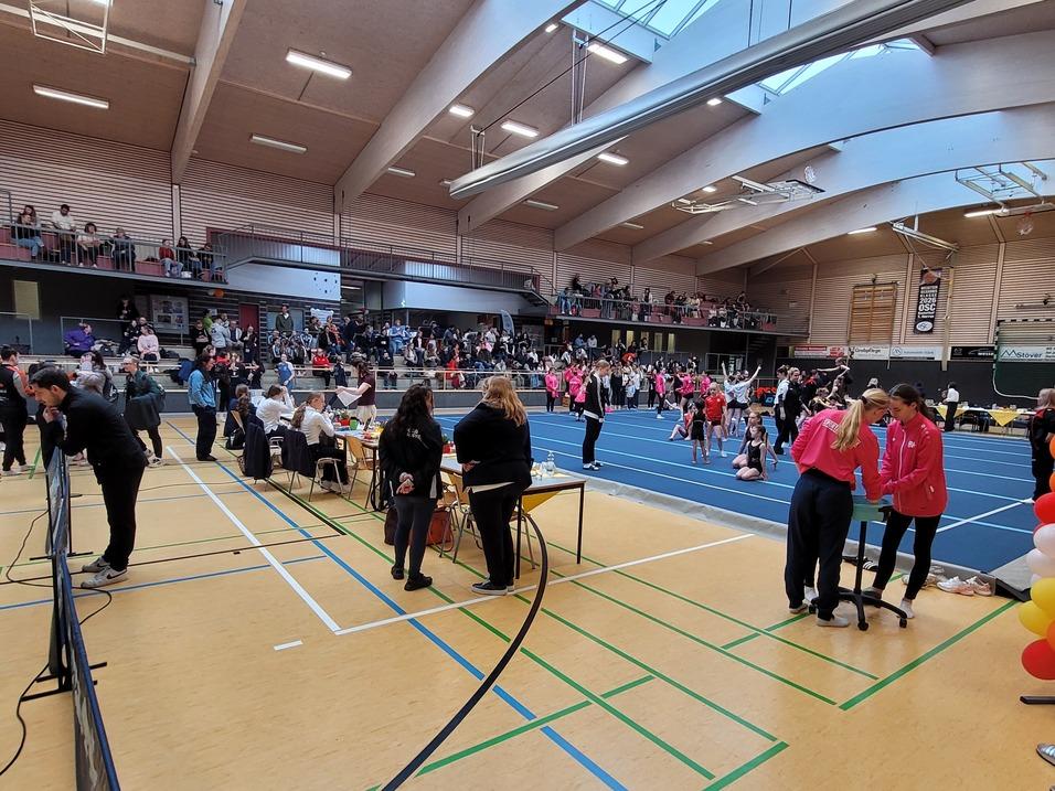 Viel belebte Sporthalle mit Zuschauern, Teilnehmern und buntem Luftballonbogen bei einem Wettkampf.
