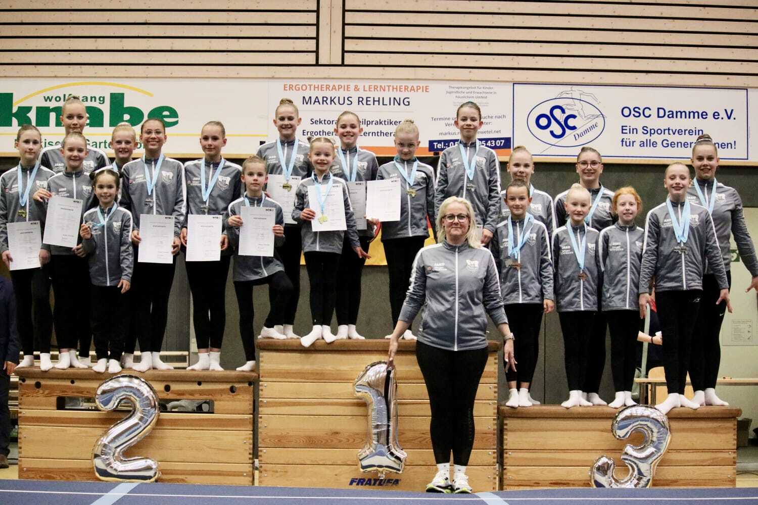 Gruppe von Turnerinnen mit Medaillen und Urkunden auf einem Treppchen, lächelnd in Sportkleidung, vor einer Wand mit Vereinslogos.