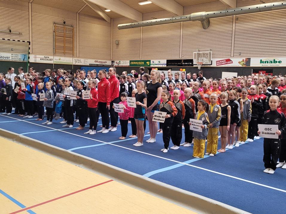 Gruppenfoto von Sportlern in Wettkampfkleidung, versammelt auf einer blauen Turnmatte, mit Namensschildern in den H&auml;nden.