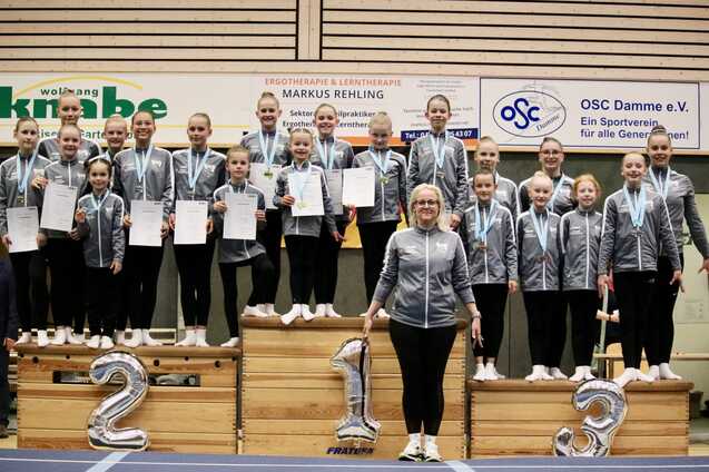 Gruppe von Turnerinnen mit Medaillen und Urkunden auf einem Treppchen, l&auml;chelnd in Sportkleidung, vor einer Wand mit Vereinslogos.
