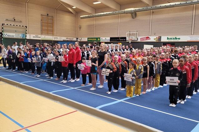 Gruppenfoto von Sportlern in Wettkampfkleidung, versammelt auf einer blauen Turnmatte, mit Namensschildern in den H&auml;nden.