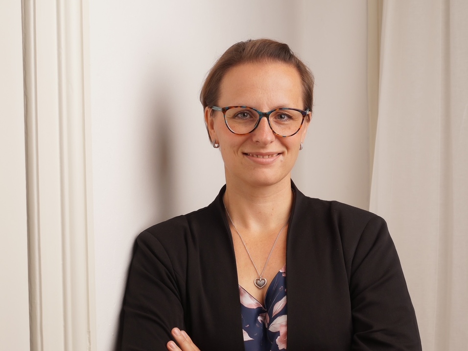 Frau mit Brille und gefalteten Armen steht an einer Wand neben einem Vorhang. Sie tr&auml;gt einen schwarzen Blazer und ein buntes Oberteil.