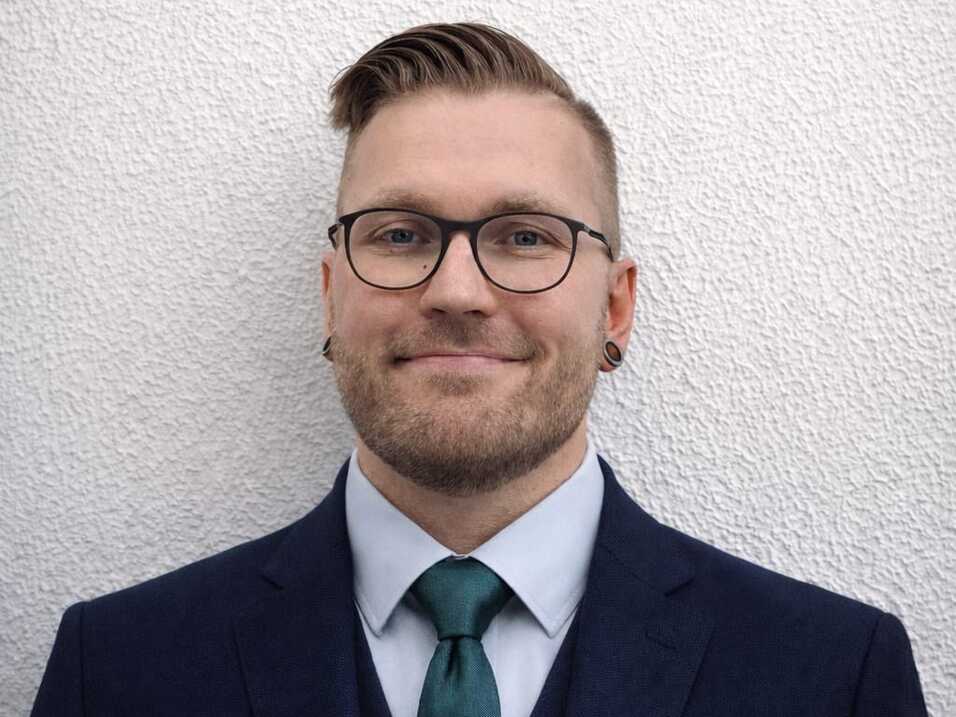 Mann mit Brille, Anzug und Krawatte steht vor einer strukturierten weißen Wand.