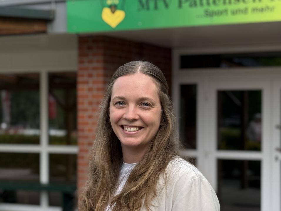 Frau mit langen Haaren lächelt vor dem Eingang des MTV Pattensen 1909 Vereinsgebäudes.