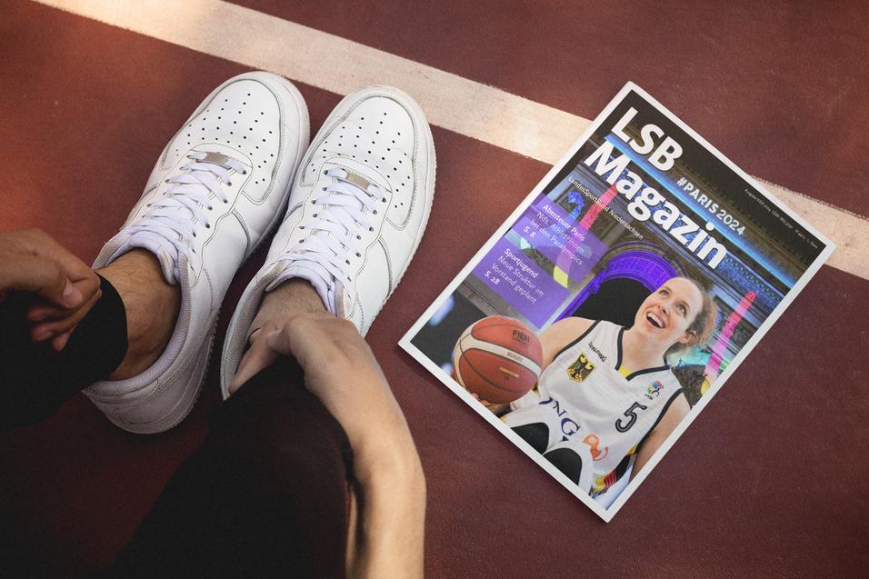 Person im Sportoutfit mit weißen Sneakers sitzt neben einem Magazin auf einem Basketballplatz.