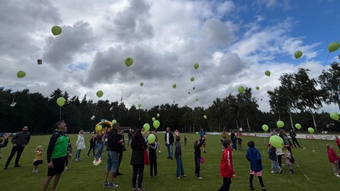 Gruppierung von Menschen auf einem Rasenplatz, die gr&uuml;ne Luftballons in den klaren Himmel steigen lassen.