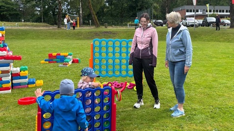Zwei Frauen stehen neben einem gro&szlig;en, bunten Spielger&auml;t, w&auml;hrend ein Kind davor spielt. Im Hintergrund ist eine Wiese zu sehen.