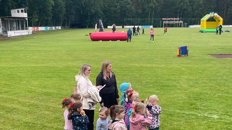 Gruppierung von Kindern und zwei Erwachsenen auf einer Wiese, umgeben von Spielger&auml;ten und einer gr&uuml;nen Landschaft.