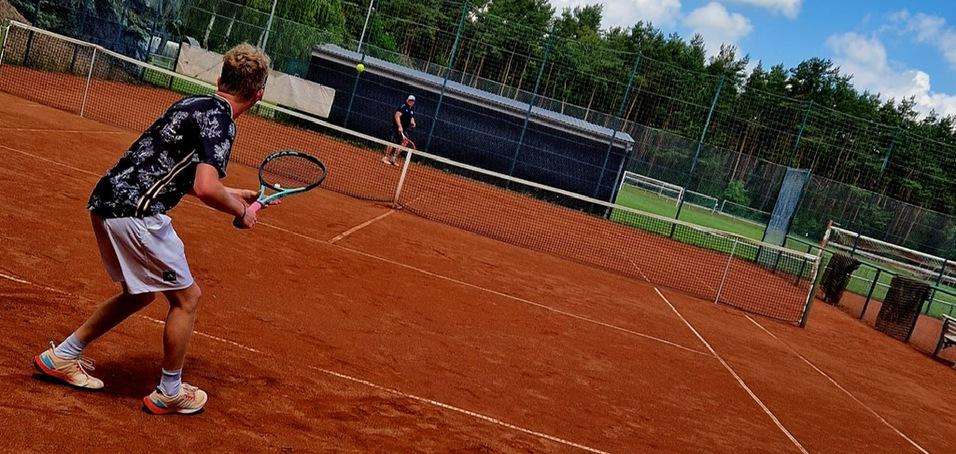 Junger Mann mit Tennisschl&auml;ger schl&auml;gt auf einem roten Sandplatz gegen einen Gegner in der Distanz. Wolken am blauen Himmel.
