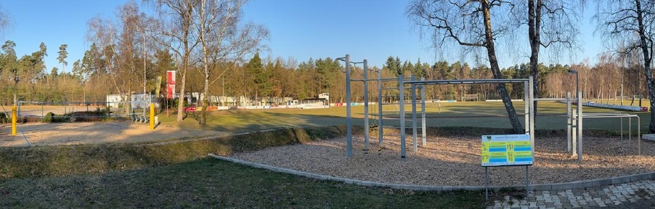 Panoramablick auf einen Spielplatz mit Schaukeln, einer Trainingsstation und einer gr&uuml;nen Wiese, umgeben von B&auml;umen.