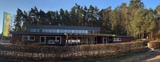 Panoramabild eines modernen Geb&auml;udes mit gro&szlig;en Fenstern, umgeben von einem Waldbereich und einem gepflegten Garten.