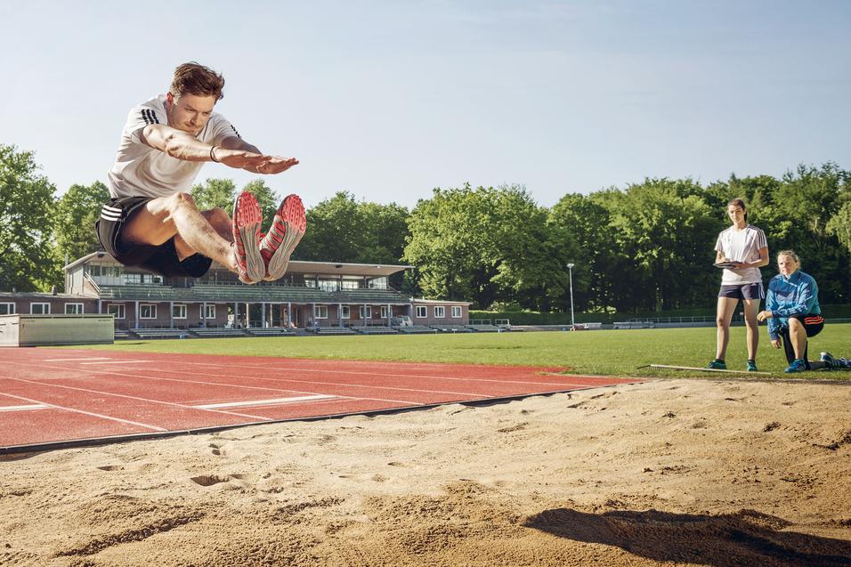 Weitspringer in der Luft beim Absprung über eine Sandgrube, daneben zwei Beobachterinnen.