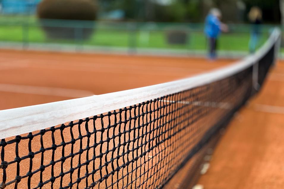 Nahaufnahme eines Tennisnetzes auf einem roten Sandplatz, Hintergrund unscharf mit grünen Bäumen.