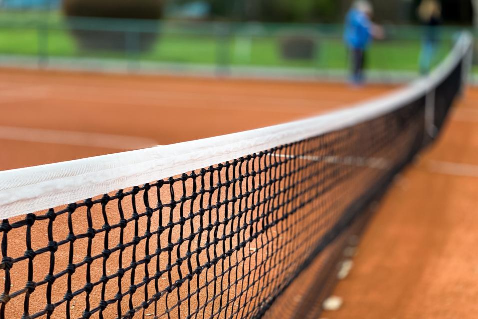 Nahaufnahme eines Tennisnetzes auf einem roten Sandplatz, Hintergrund unscharf mit grünen Bäumen.